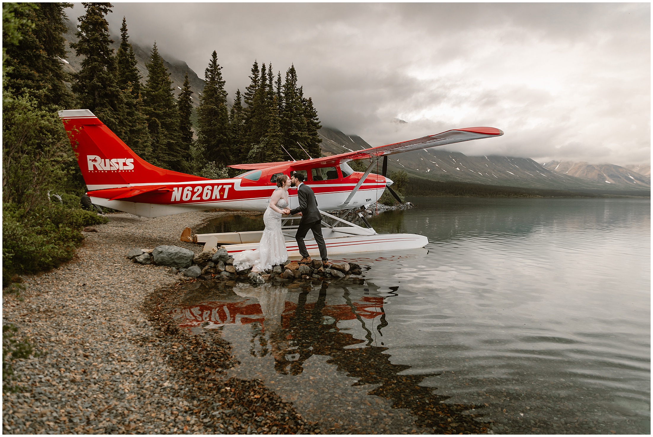 Newlyweds kiss on the rocks beside their beached red floatplane — a floatplane elopement in Alaska