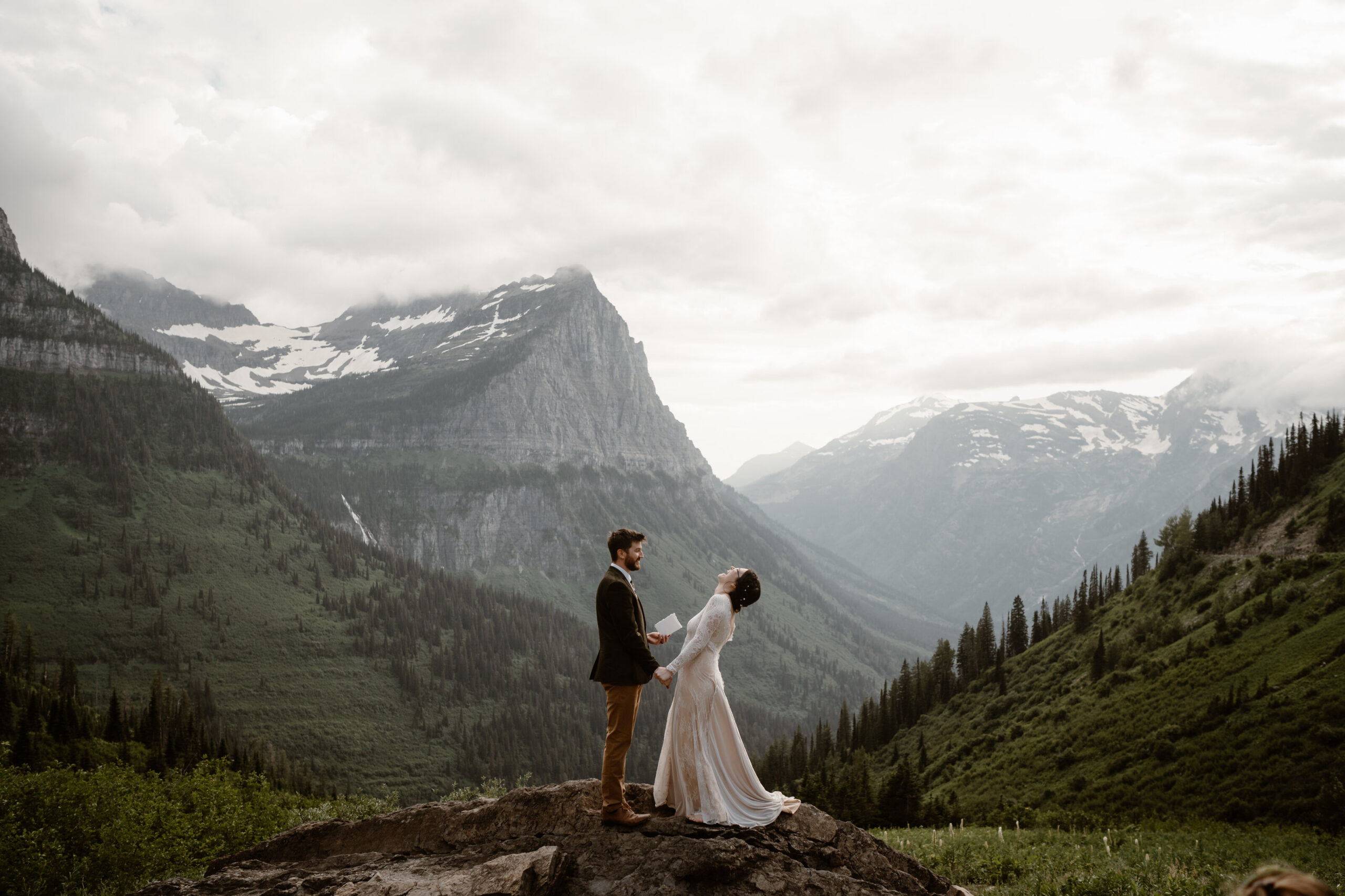 Bride leans back laughing while her groom reads vows on a rock above a sweeping valley during their Glacier National Park elopement